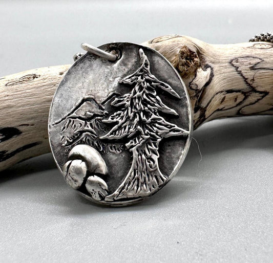 Silver pendant with a redwood tree, boulders, and mountains scribed on it.