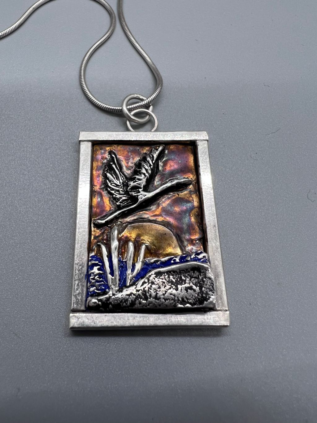 Silver framed PMC silver clay pendant of a crane flying over the sunrise at a pond with colored surfaces.