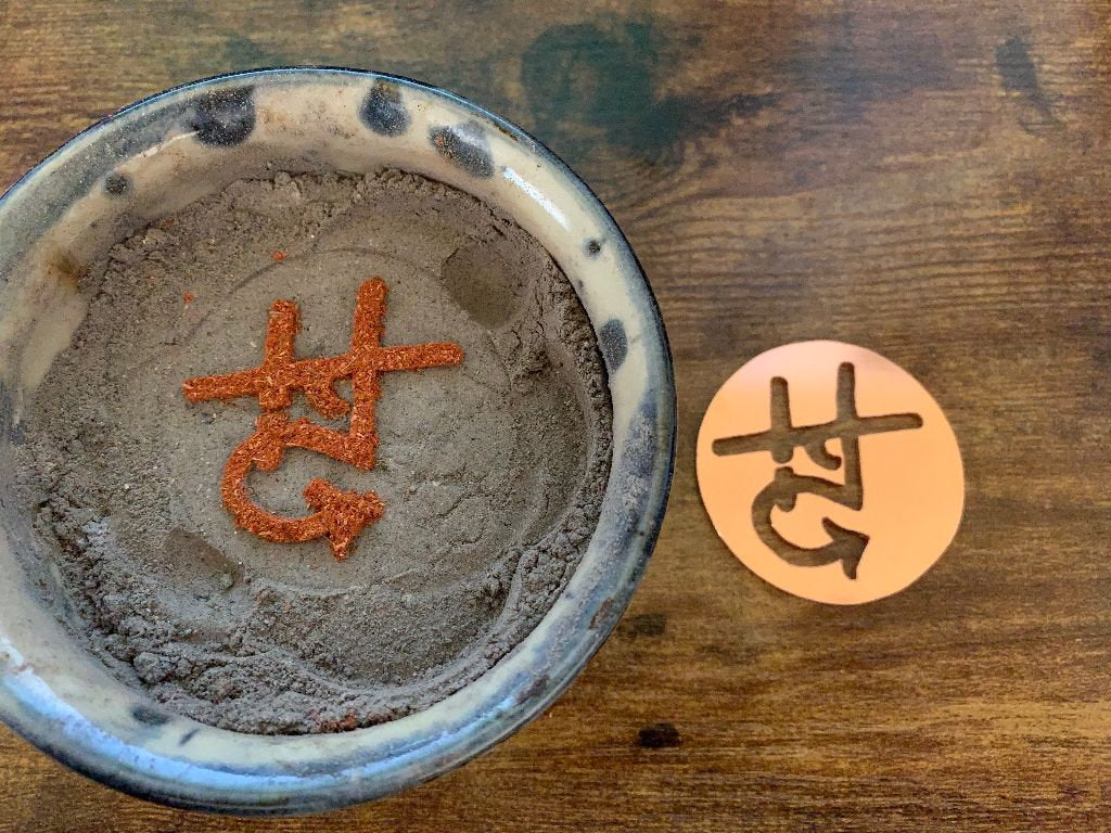 Round copper piece with saw cut logo and incense trail on ash.